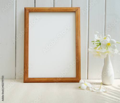 Mock up poster wooden frame and beautiful Frangipani tropical flower in modern white vase on beige table and wood wall background with palm leaf shadow