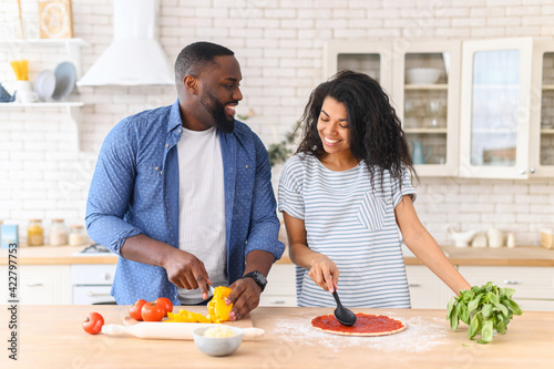 Romantic couple is cooking on kitchen. Handsome man cuts vegetables and attractive young woman prepares the dough by spreading it with tomato paste, for dinner there will be an appetizing little pizza