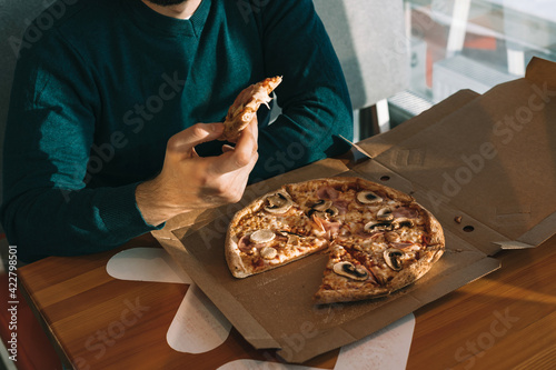 The hand of a man. Handsome bearded European man in a green sweater eats pizza with ham and mushrooms in a cardboard box and drinks a drink in a paper glass on a wooden table in a cafe.