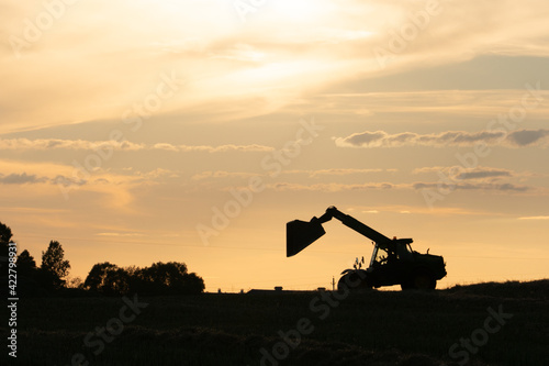 Silhouette of a working excavator with a raised bucket against the background of sunset and clouds. The field is operated by agricultural machinery. The season of harvesting or planting the crop.