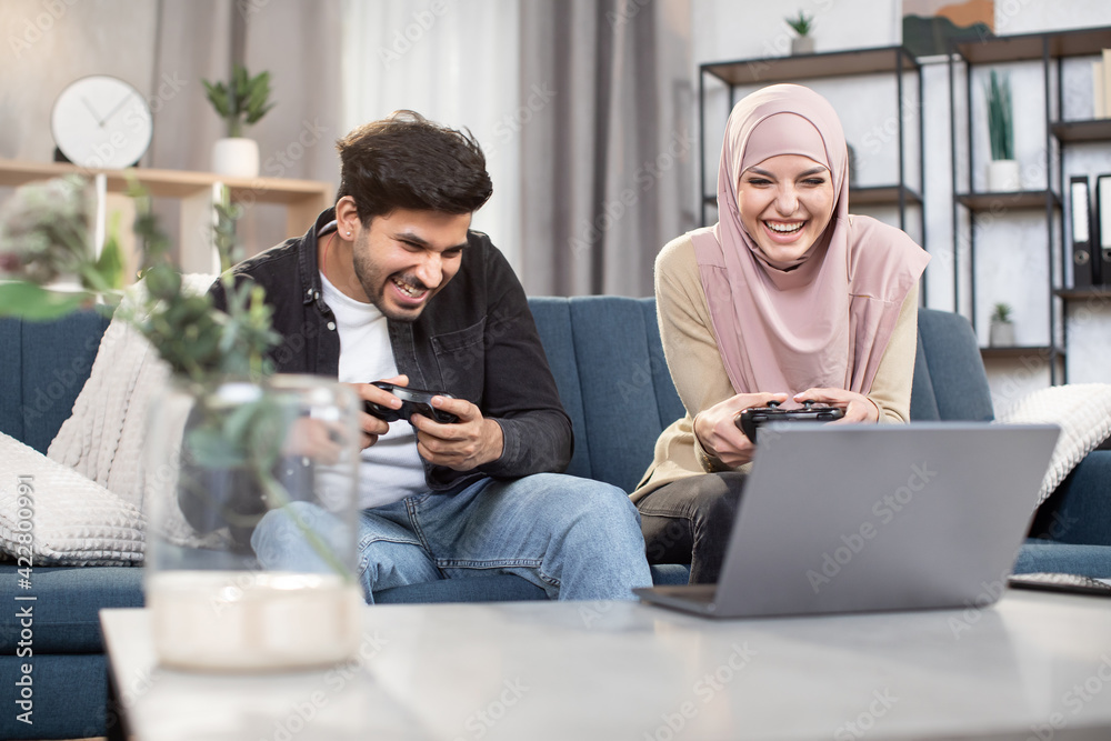Laughing 30-aged funny Muslim couple having fun together while playing video games at home using gamepads. Family leisure, playing video games. Laughing 30-aged funny Muslim couple having fun together while playing video games at home using gamepads. Family leisure, playing video games.