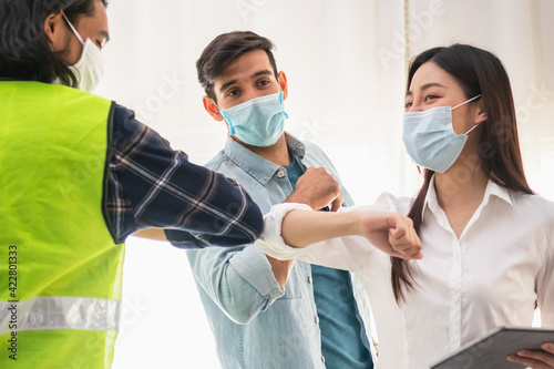 Group of smiling young caucasian, asian colleagues or contractors, engineer wearing face masks and bumping elbows to protection at workplace while coronavirus is pandemic.Greeting in Covid concept,