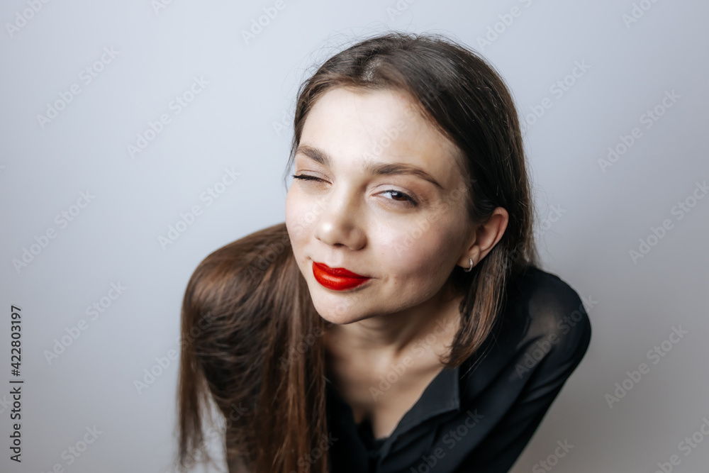 A beautiful girl with red lips winks. Large portrait on a light background A beautiful girl with red lips winks. Large portrait on a light background