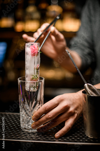 bartender holds piece of ice with tongs and puts it into crystal glass on bar counter