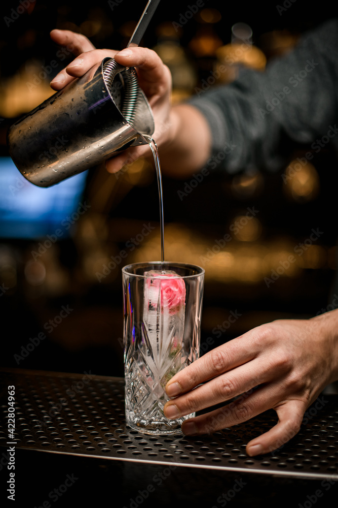 close-up of crystal glass with ice in which bartender pours drink from steel mixing cup