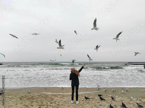 A girl with long hair standing on the beach and feeding seagulls with bread with a beautiful sea background. Girl feeds seagulls and pigeons at the seashore. 