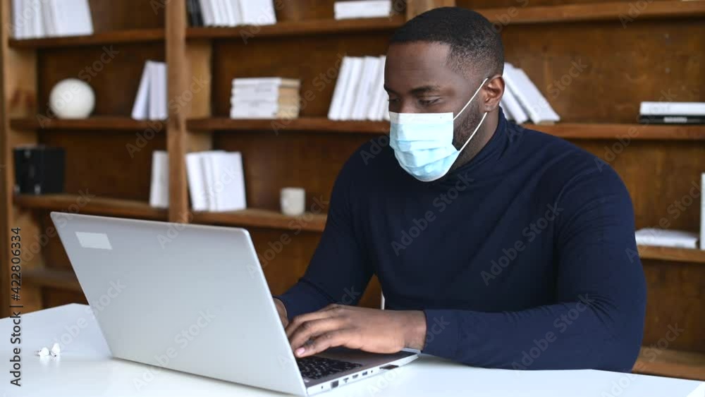 Focused African-American wearing medical mask working with a laptop in contemporary office space with bookshelves on the background, social distancing and protective measures for office employees Focused African-American wearing medical mask working with a laptop in contemporary office space with bookshelves on the background, social distancing and protective measures for office employees