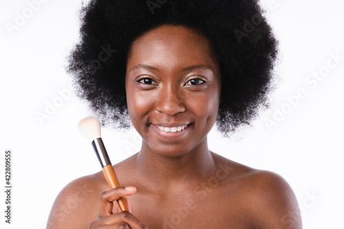 Smiling young african girl in bath towel holding make-up brush isolated in white background