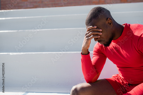 Fotografi Athletic black man in sportswear sitting on stairways cover face with hand and look down