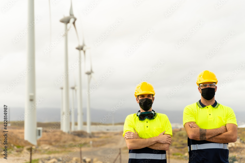 Front view of two skilled workers in a wind turbine power station. Two male electrician workers in reflective vests and hard hats. Clean energy, renewable energy and environment concept.