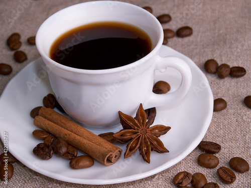 A white cup of espresso stands on a burlap table with scattered coffee beans, a cinnamon stick and stars of anise.