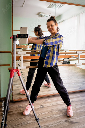 A fitness trainer leads an online class from a hall with mirrors, a woman demonstrates a warm-up exercise in front of a smartphone mounted on a tripod. 