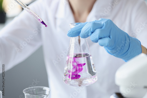 Scientist in laboratory holds transparent flask of liquid and digs purple reagent into it photo