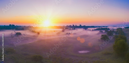 Beautiful rural landscape with foggy colorful trees, orange sky with sun. Aerial view of village in fog with golden sunbeams at sunrise.