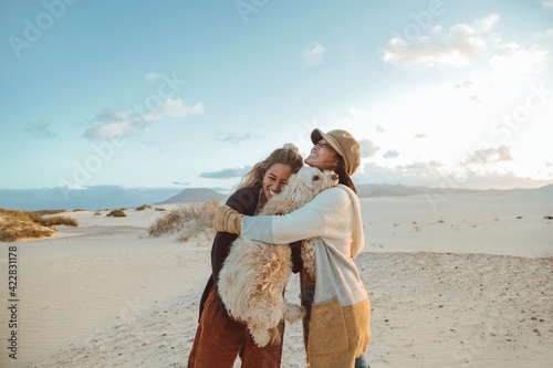 Two beautiful filipino women laughing and hugging each other and their dog - Friends having a great time together walking in a desert