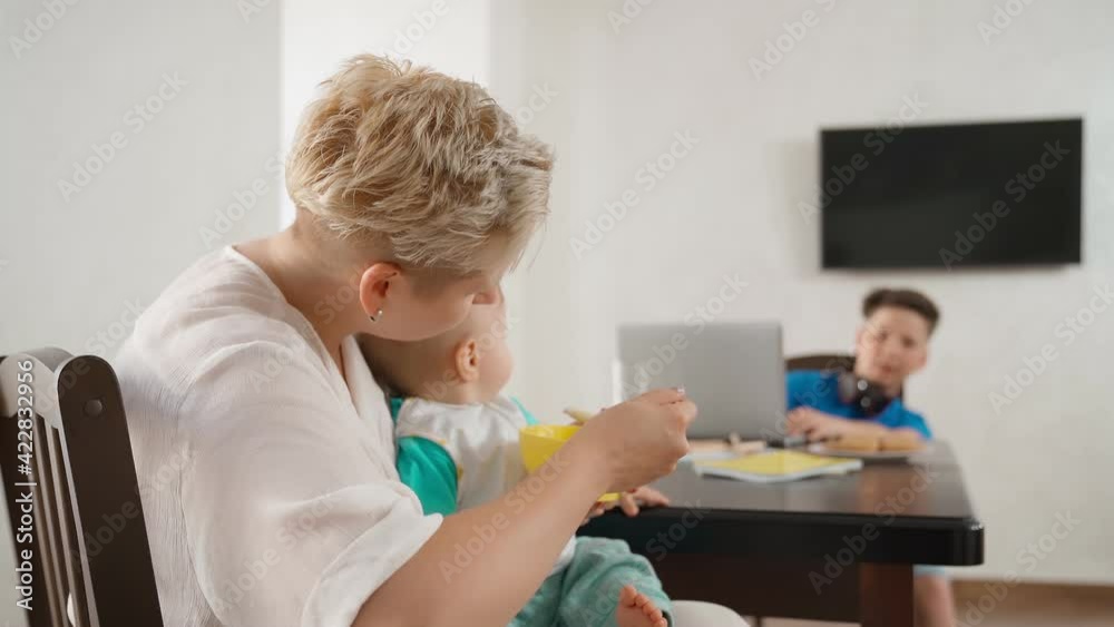 Mother spoon feeding baby boy while teenager using laptop Mother spoon feeding baby boy while teenager using laptop