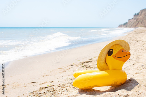 Yellow inflatable duck ring in summer straw hat laying on sandy empty beach near blue wavy ocean in sunny day. Protection swim tube for kid. Summer travel vacation resort concept. Copy space