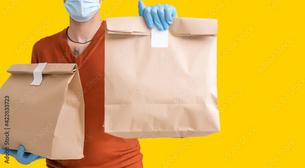 Delivery man employee in mask glove hold craft paper packet with food isolated on yellow background studio Service quarantine pandemic coronavirus virus 2019-ncov concept Delivery man employee in mask glove hold craft paper packet with food isolated on yellow background studio Service quarantine pandemic coronavirus virus 2019-ncov concept