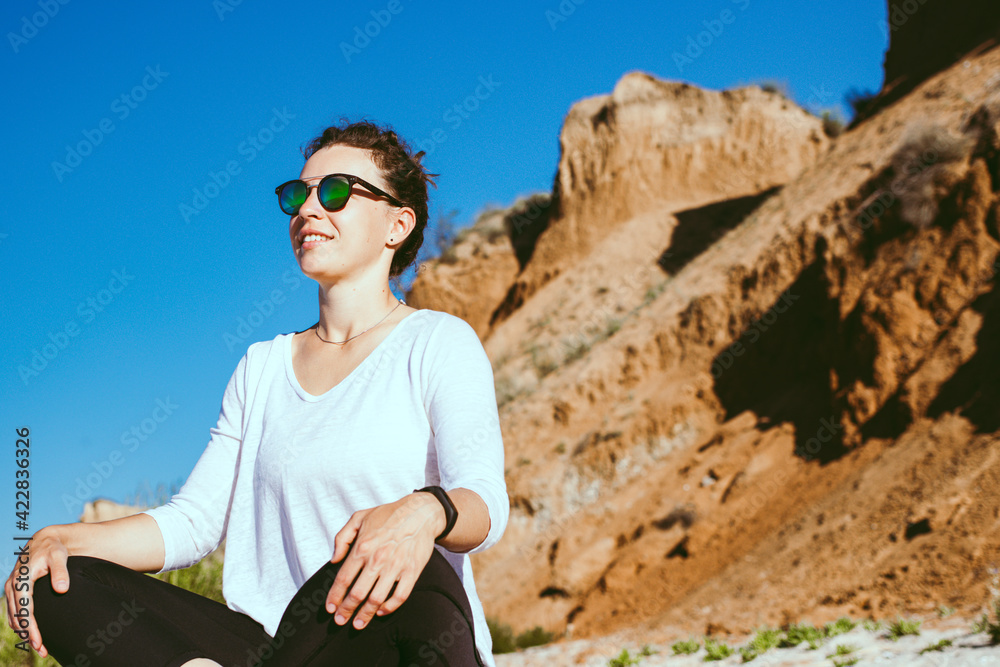 Happy young woman practicing breathing yoga, sitting in lotus pose on sandy brown cliff and blue sky background. Smiling female athlete meditating outdoor in sunny day,enjoying outdoor training class