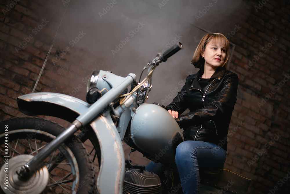 Obraz premium Young biker woman is sitting on the retro motorbike in the old garage.