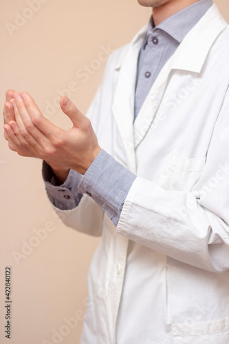 A young man in a white coat raised his hands up and pray. Beige background. Doctor praying. A Muslim makes dua.