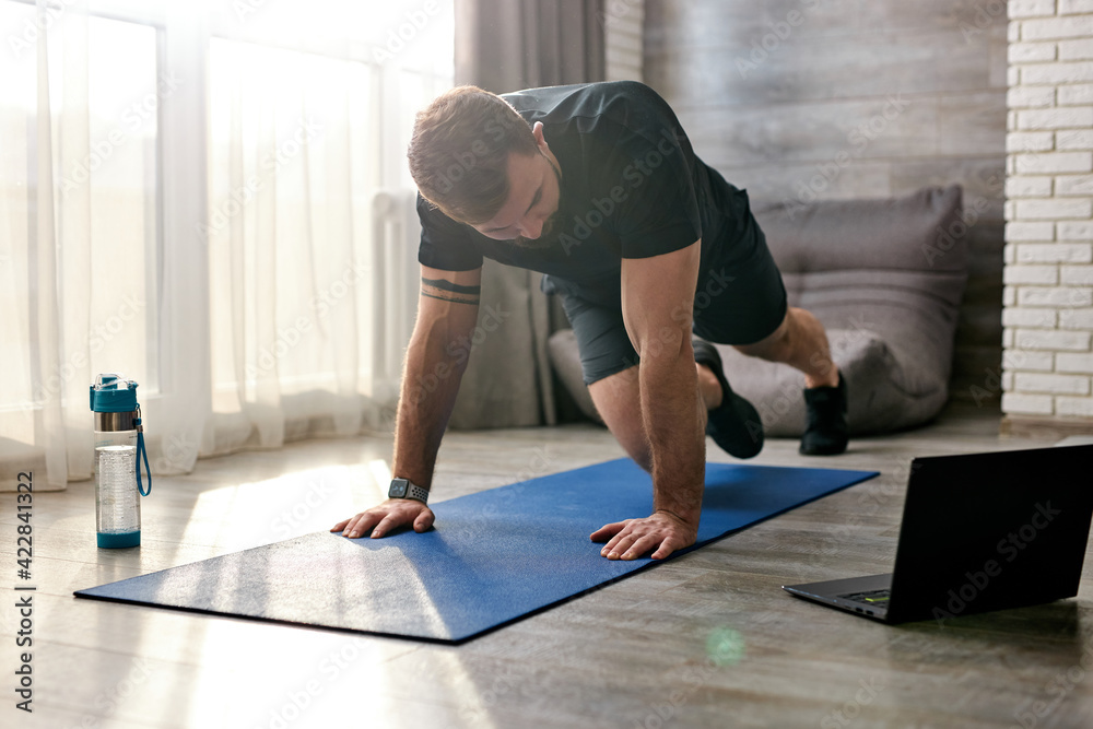 Sportive male doing exercises on mat watching online training on laptop