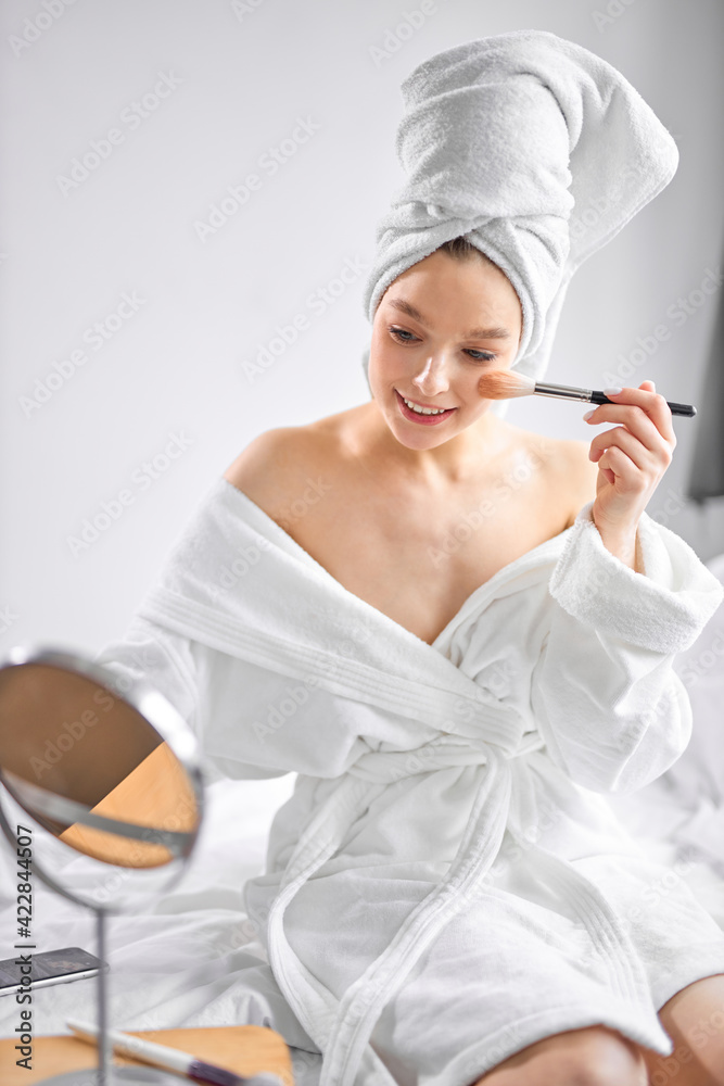 Cute Caucasian woman using makeup brush and smiling in front of small mirror Cute Caucasian woman using makeup brush and smiling in front of small mirror