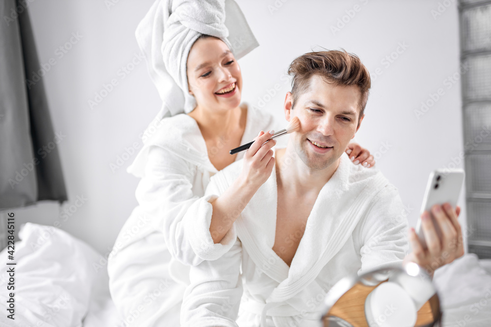 cheerful young caucasian couple in bathrobes and towel have fun, woman doing make-up on boyfriend's face cheerful young caucasian couple in bathrobes and towel have fun, woman doing make-up on boyfriend's face