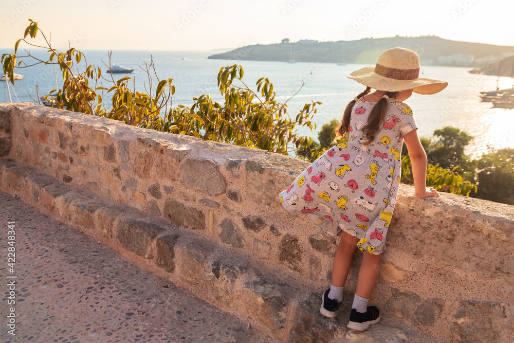 Obraz premium A little girl with a big hat looks at the sea and the city. A girl in the castle of Bodrum, Turkey. The baby is resting and going to the sights.