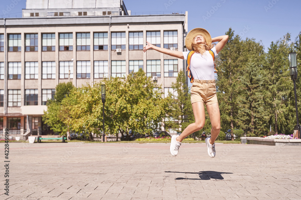Obraz premium Happy young woman celebrate end of lockdown. Borders are open. Jumping at street if town. Summer time. Staycation concept. Female tourist. Backpack, hat, t-shirt, shorts. Sunny day