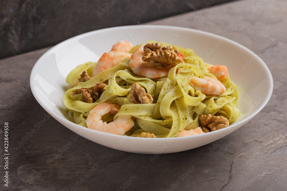 Dish of Tagliatelle pasta with shrimps, pesto sauce, walnut on white plate on dark stone table. Mediterranean cuisine.