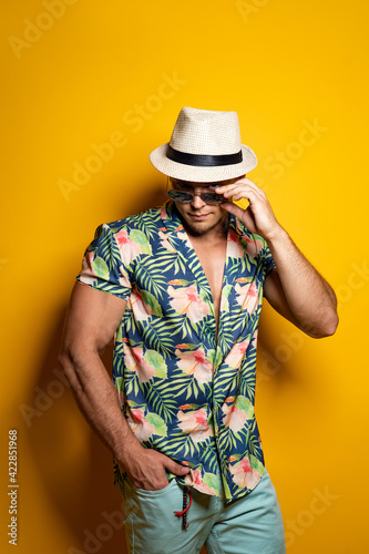 Young handsome Caucasian man wearing Hawaiian shirt, hat, sunglasses posing against yellow wall