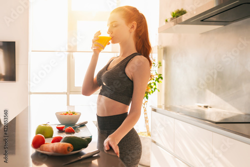 Athletic young red haired woman in the kitchen drinks a glass of fruit centrifuged juice