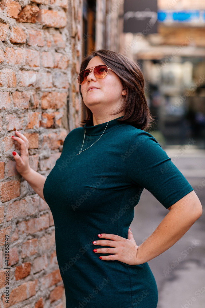 Portrait of stylish young pretty hipster woman in sunglasses and green dress, vintage urban background, street photo brick wall, looking away, Happy beautiful girl, body positivity, casual clothes