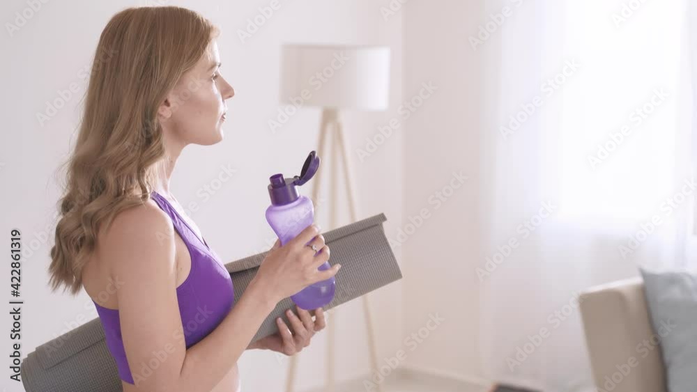 Portrait of smiling cute young woman with sports mat in hand drinks water after workout at home. Sunlight, side view Portrait of smiling cute young woman with sports mat in hand drinks water after workout at home. Sunlight, side view