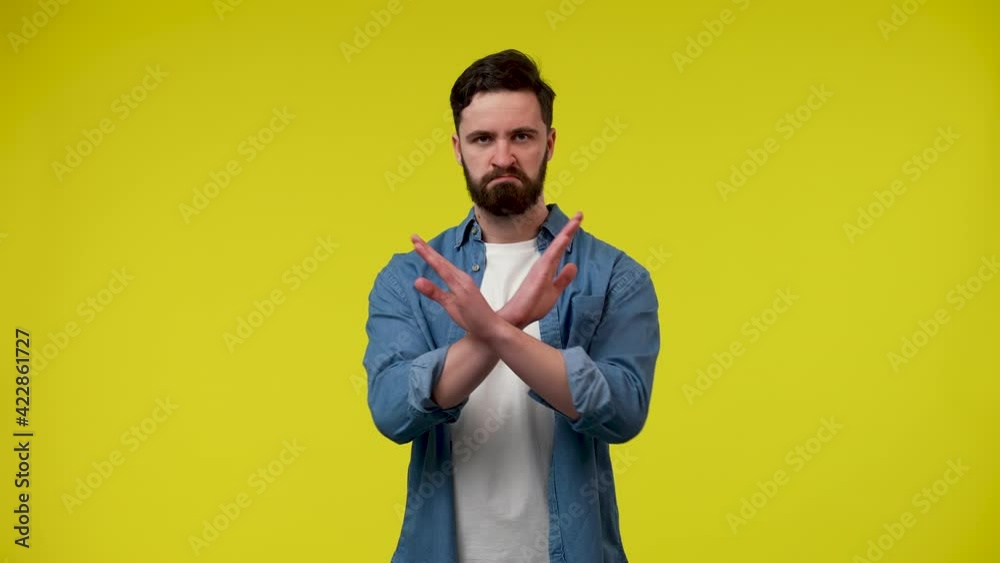 A disgruntled bearded man says no with a stop sign, extending his arms forward, then crosses them and nods his head. A man in the studio on a yellow background. Close up. Slow motion ready 59.97fps.