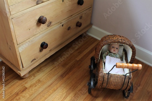 Tablou pe pânză Small woven baby carriage with fake baby inside a play room with wooden floors and cabinets