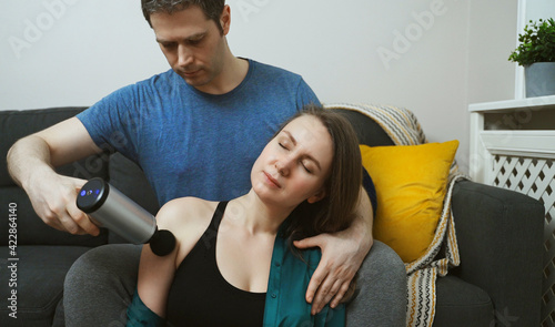 Man massaging woman with massage percussion device at home.