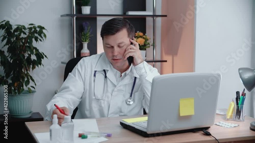 Wallpaper Mural A handsome male doctor sits at a table talking on the phone writes down the data that he is told on a sheet of paper. Torontodigital.ca