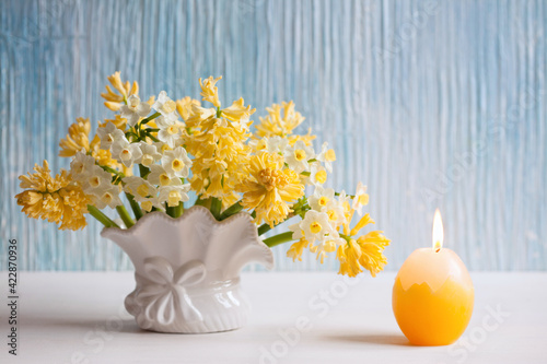 Fototapeta Naklejka Na Ścianę i Meble -  Easter background card with yellow flowers hyacinths, daffodils in a vase and a candle in the shape of an egg on the table.