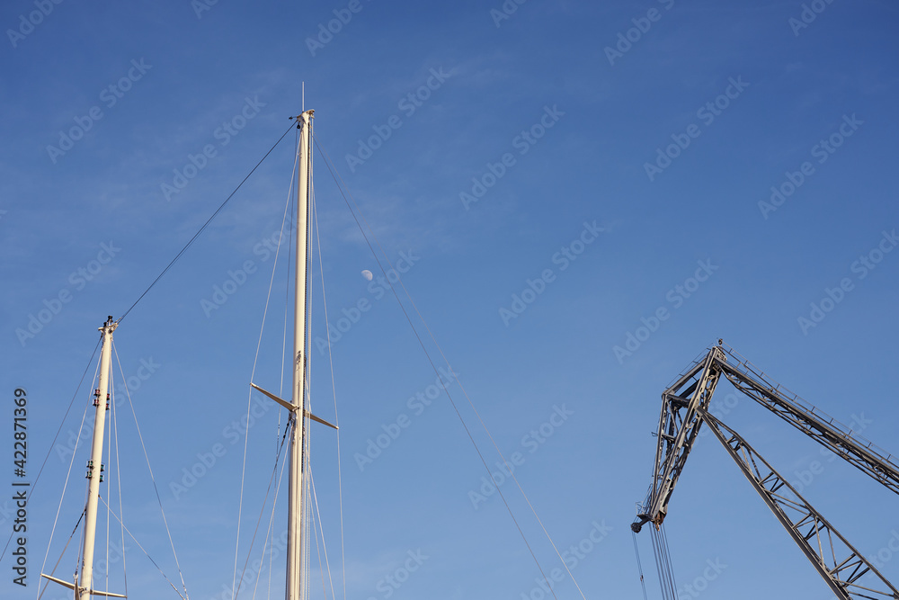 The yacht's masts and an old crane against the blue sky with the moon.