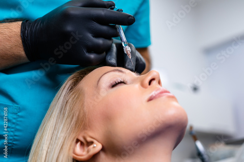 Attractive young woman is getting a rejuvenating injections. She is sitting calmly at clinic. The expert beautician is filling female lips by hyaluronic acid.