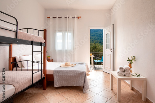 White modern interior of family bedroom with classic double wooden bed and bunk bed with metal stairs, open door to summer olive garden view balcony with elegant terrace furniture