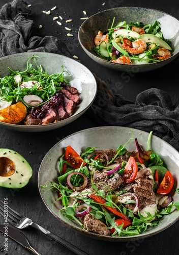 Fresh salads with avocado, shrimps, beef steak, tomatoes, green leaves, arugula, spinach, chicken breast and sweet sauce on dark wooden background. Healthy food, clean eating, closeup