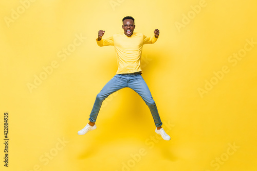 Young cheerful excited African man jumping and screaming with clenching hands on isolated yellow studio background