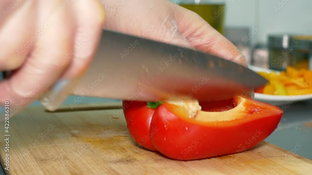In the restaurant kitchen, a red bell pepper is cut into two halves on a wooden kitchen board, the core is cut out, and the work is performed wearing disposable plastic gloves. Close-up. In the restaurant kitchen, a red bell pepper is cut into two halves on a wooden kitchen board, the core is cut out, and the work is performed wearing disposable plastic gloves. Close-up.
