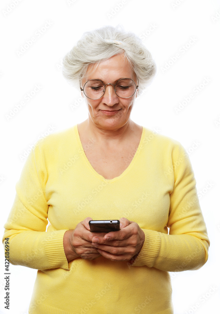 Fototapeta premium Lovely senior lady in casual outfit cheerfully smiling and looking at camera while standing on white background and using modern smartphone
