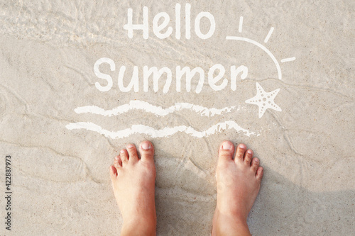 Hello Summer concept, kids foot standing on the clean white sandy beach with soft wave, Hello Summer wording, sun and starfish in white color on the sand background