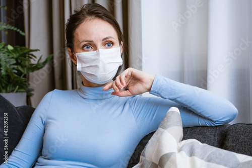 Woman in a protective mask on the couch looks to the side with a pensive look