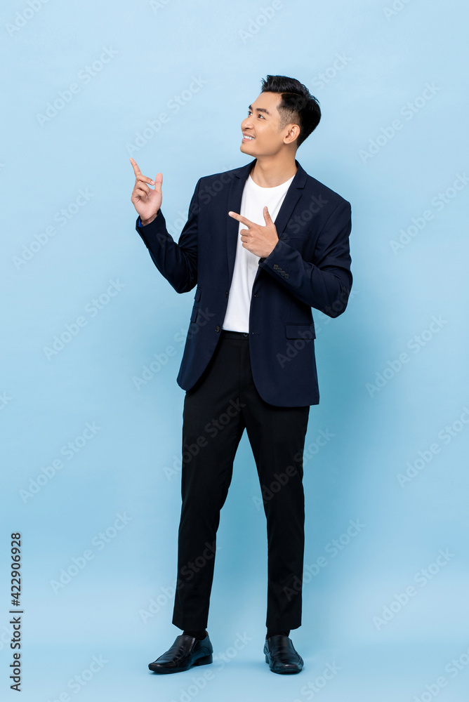 Full length portrait of happy smiling young handsome southeast Asian businessman pointing and looking upward on light blue studio background